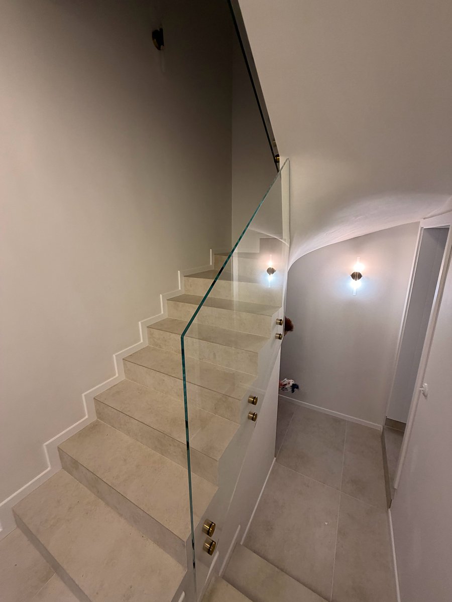 Schody wyłożone jasnymi płytkami gresowymi ze szklaną balustradą, gładkie ściany i kinkiety